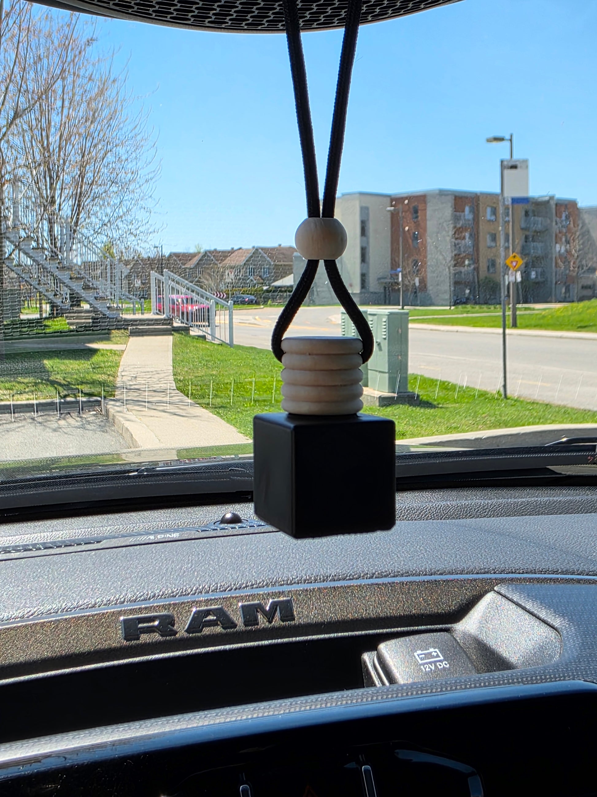 Imperial Ancient Imperial Oud Car Air Freshener – Square black bottle with wood bead cap, hanging from rearview mirror inside a vehicle, with sunny outdoor background.